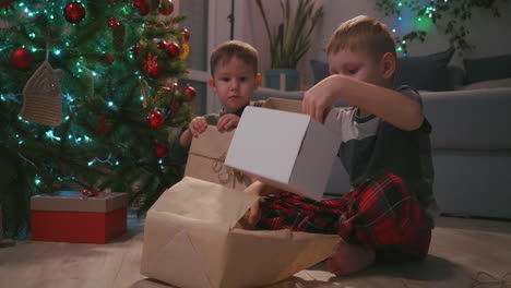 Zwei-Jungen-öffnen-Weihnachtsgeschenke-Und-Sitzen-Unter-Einem-Geschmückten-Weihnachtsbaum-Inmitten-Von-Girlanden.-Hochwertiges-4K-Filmmaterial